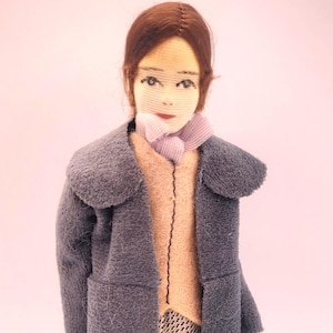 May include: A vintage doll dressed in a gray coat with a light pink sweater and patterned pants. The doll has brown hair and a light pink scarf. The doll is wearing black shoes. The doll is standing on a white surface.