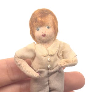 May include: A small antique doll with a fabric body, wearing a cream-colored shirt, gray shorts, and brown shoes. The doll has red hair and is approximately 4 inches tall. The doll's arms are slightly outstretched.