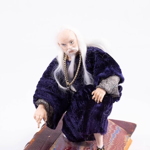 May include: A white-haired, bearded figurine wearing a purple robe and a gold chain necklace sits on an antique book with a marbled cover.