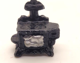 May include: A miniature black stove with a detailed design. The stove has a small chimney, a round side piece, and a silver-colored door. The stove is likely a decorative item or part of a dollhouse set.