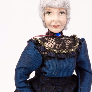 Op de afbeelding: Een porseleinen pop met grijs haar, gekleed in een blauwe jurk met zwarte kantversiering en een blauwe juwelenketting.