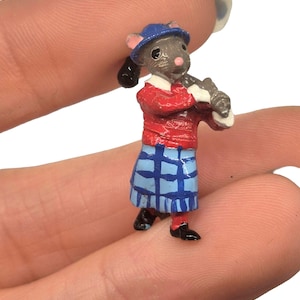 May include: Miniature figurine of a mouse dressed in a red sweater, blue plaid skirt, and a blue hat. The mouse is holding a musical instrument. The figurine has black shoes and ears. The background is white.