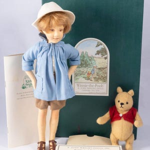 May include: A set of Winnie-the-Pooh dolls featuring Christopher Robin in a blue coat and a small Pooh bear in a red vest. The collection includes a certificate of authenticity and a green box with the Winnie-the-Pooh logo.