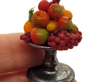 Puede incluir: Arreglo de frutas en miniatura en un cuenco con pedestal plateado. El arreglo incluye frutas rojas, naranjas y verdes, como manzanas, uvas y cítricos. El cuenco tiene una base redonda y un tallo. La fruta está dispuesta de forma decorativa.