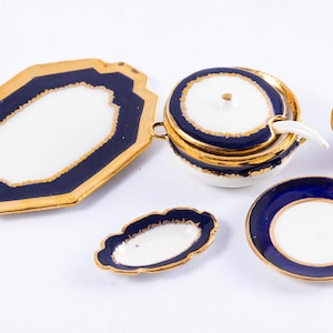 May include: A collection of antique porcelain serving pieces. The set includes a large serving tray, a covered bowl with a spoon, a small creamer, a small oval dish, and a small plate. Each piece has a white interior, a navy blue border, and gold trim.