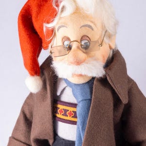 May include: A handmade doll depicting an elderly man wearing a red Santa hat with a white bobble, gold-rimmed spectacles, and a white moustache. He is dressed in a brown coat, a blue tie, and a sweater with a decorative band.