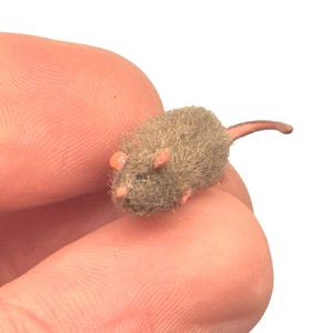 May include: A miniature, fuzzy mouse figurine in shades of brown and tan, with pink ears and a tail. The tiny rodent is held between two fingers, showcasing its small size and detailed craftsmanship. The mouse is a decorative item.