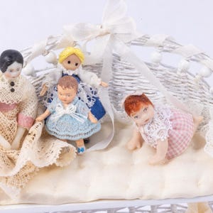 May include: A white wicker basket with four antique porcelain dolls. The dolls are dressed in vintage clothing, including lace and blue dresses. One doll is crawling, and the others are standing. The basket is decorated with a white ribbon.