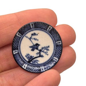 May include: A small, round, blue and white decorative plate. The plate features a bird perched on a branch, with a blue and white patterned border. The plate is held in a hand.