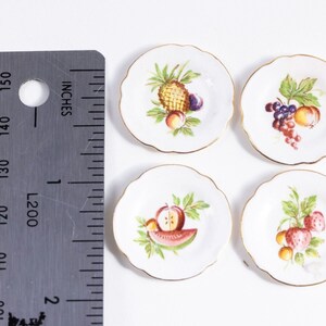 K&ouml;nnte beinhalten: Set aus vier kleinen wei&szlig;en Keramiktellern mit goldener Verzierung. Jeder Teller zeigt ein anderes Fruchtdesign: Ananas, Trauben, Wassermelone und Erdbeeren.