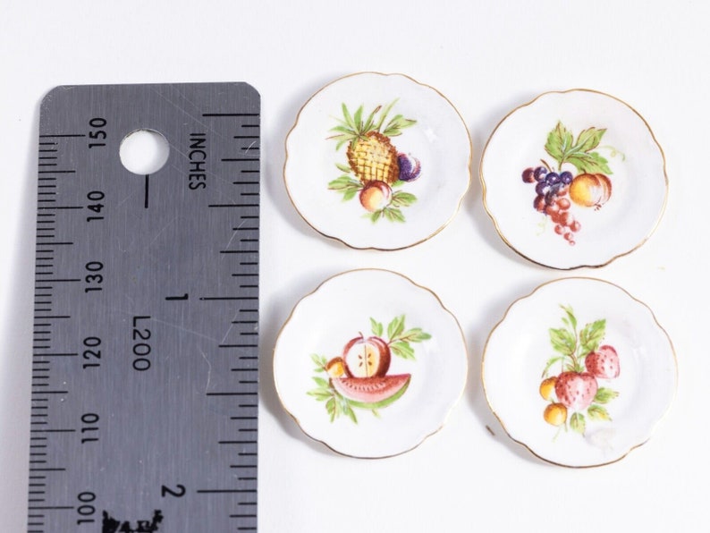 K&ouml;nnte beinhalten: Set aus vier kleinen wei&szlig;en Keramiktellern mit goldener Verzierung. Jeder Teller zeigt ein anderes Fruchtdesign: Ananas, Trauben, Wassermelone und Erdbeeren.