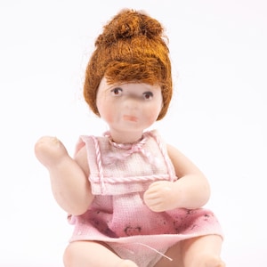 May include: A porcelain doll with red hair, wearing a pink dress with white trim. The doll is sitting with its legs crossed and its arms bent.