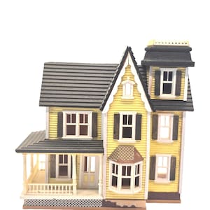 Puede incluir: Una casa victoriana amarilla en miniatura con techo gris, contraventanas negras y porche. La casa tiene múltiples ventanas y un diseño detallado, mostrando un estilo arquitectónico clásico. Es un artículo decorativo.