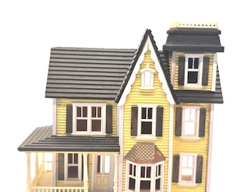 Könnte beinhalten: Ein gelbes viktorianisches Miniaturhaus mit grauem Dach, schwarzen Fensterläden und einer Veranda. Das Haus hat mehrere Fenster und ein detailliertes Design, das einen klassischen Architekturstil zeigt. Es ist ein Dekorationsartikel.