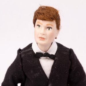 May include: A miniature doll figure of a man in a black suit with a white collared shirt and a black bow tie. The doll has red hair and green eyes. The figure is a collectible item.