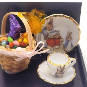 Puede incluir: Mini cesta de Pascua con un lazo rosa, llena de caramelos coloridos y un chocolate envuelto en papel de aluminio morado. Incluye un juego de taza y platillo pequeños, y un plato decorativo, todos con ilustraciones de conejos.