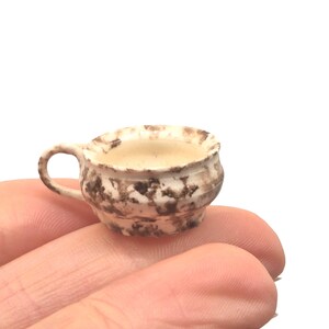 May include: A miniature ceramic teacup with a handle, featuring a white base with brown mottled patterns. The cup is filled with a light-colored liquid, possibly tea or coffee. The cup is held in a person's hand.