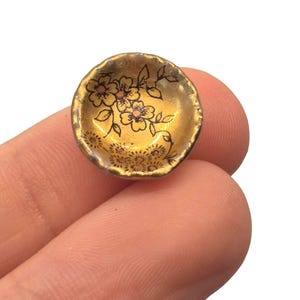 May include: A small, gold-colored bowl with a floral design. The bowl has a slightly irregular rim and is decorated with black line art flowers and pink accents. The bowl is held between two fingers.