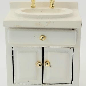 May include: Miniature white bathroom vanity with a sink and two gold-colored faucets. The vanity has a drawer and two cabinet doors, each with a gold knob. A charming piece for a dollhouse or miniature collection.