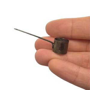 以下が含まれることがあります： 長い細い柄が付いた暗い金属製のミニチュア計量カップ。小さな円筒形の容器は、風化したダークグレーの仕上げが特徴で、側面から伸びる細く真っ直ぐなアームの先端には小さな穴が開いています。