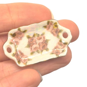 Puede incluir: Una pequeña bandeja rectangular de cerámica con un diseño floral. La bandeja presenta una base de color crema con rosas rosas y hojas verdes. Tiene dos pequeños agujeros en cada extremo y un borde dorado.