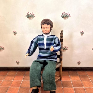 May include: A miniature doll with brown hair and a beard, wearing a blue and green striped long-sleeve shirt with a white collar and solid green trousers. The doll is seated on a small wooden chair with a woven seat, featuring dark brown shoes.