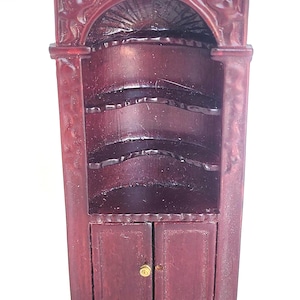 May include: A small, ornate, burgundy-colored cabinet with a decorative arched top and two lower doors. The cabinet has multiple shelves and a gold-colored knob on the doors. The cabinet is made of wood and has a vintage appearance.