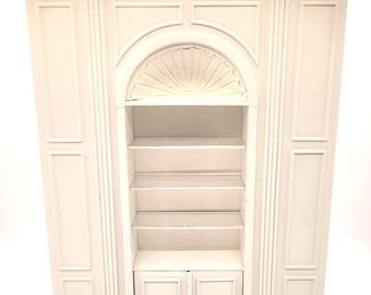 May include: A white, ornate bookshelf with an arched center section and multiple shelves. The bookshelf features decorative molding and a cabinet with two doors at the bottom. The overall design is classic and elegant.