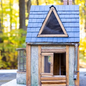 May include: A miniature wooden house with a blue shingled roof and a triangular window. The house has a rustic appearance with wooden siding, a small window, and a chimney. The background is a blurred view of a forest.