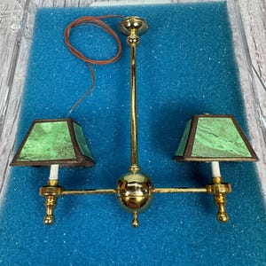 Può includere: Un lampadario dorato a due luci con paralumi in vetro verde. Il lampadario è in una scatola di plastica trasparente con imbottitura in schiuma blu.