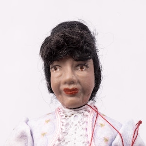 May include: A doll with a painted face, dark hair, and red lips. The doll is wearing a light purple dress with gold accents and a white lace collar. Red thread is visible near the doll's chest.
