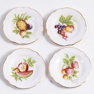 K&ouml;nnte beinhalten: Set aus vier wei&szlig;en Desserttellern aus Keramik mit goldfarbenem Rand. Jeder Teller zeigt ein anderes Fruchtdesign: Ananas, Trauben, Wassermelone und Erdbeeren.