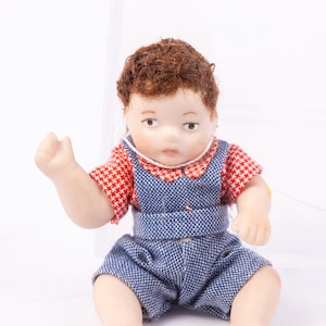 May include: A porcelain doll of a baby boy wearing a red and white checkered shirt and blue denim overalls. The doll has brown hair and is sitting with its arms outstretched.