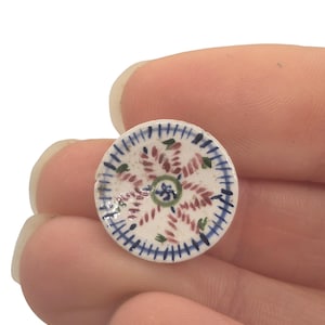Könnte beinhalten: Ein kleiner, runder, handbemalter Keramikteller mit weißer Basis. Der Teller hat ein zentrales Blumendesign in Grün und ein umlaufendes Muster aus roten und blauen Akzenten. Der Rand ist mit blauen Strichen versehen.