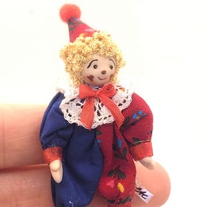 May include: A small clown doll in a red and blue costume. The doll features a red hat with a pom-pom, curly blonde hair, and a white lace collar. The outfit is split, with one side blue and the other red with floral patterns. The clown wears black shoes.