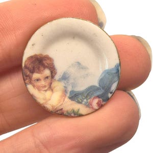 May include: A miniature decorative plate with a gold rim. The plate features a cherub design with a soft color palette of cream, blue, and red. The cherub has brown hair and is surrounded by floral and cloud-like elements.
