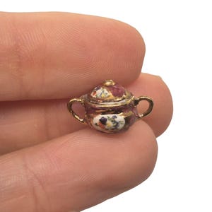 May include: Miniature decorative lidded pot with gold-colored handles and trim. The pot features a maroon base with colorful floral and abstract patterns. The pot is held between two fingers.