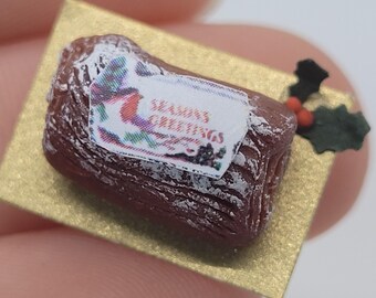 May include: Miniature Christmas Yule log cake with white frosting and a "SEASON'S GREETINGS" label. The cake sits on a gold-colored rectangular base, with a sprig of holly with red berries.