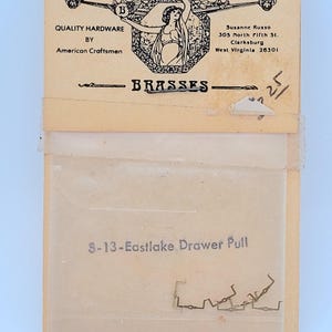 May include: A vintage package of brass drawer pulls. The package includes a paper backing with the text "QUALITY HARDWARE BY American Craftsmen" and "BRASSES". The clear plastic bag contains several small, brass-colored drawer pulls. The text "S-13-Eastlake Drawer Pull" is printed on the bag.