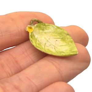 May include: A small, hand-painted leaf pendant with a yellow flower accent. The leaf is primarily green with white highlights and detailed veining. The pendant is held in a person's hand.