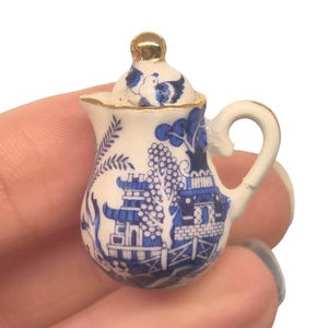 May include: Miniature white and blue ceramic teapot with a gold handle and lid. The teapot features a classic blue and white Willow pattern design. The lid has a gold knob. The teapot is held in a hand.