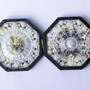 May include: An open, hinged, black octagonal box with a glass top, filled with seashells, small flowers, and decorative elements. One side features a floral arrangement. The other side has the words "BE MY LOVE" around a seashell.