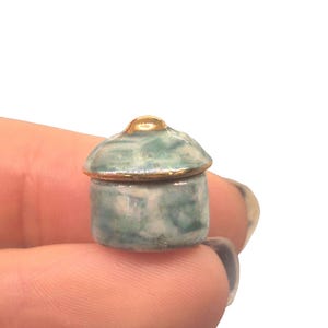 Puede incluir: Pequeño tarro de cerámica con tapa. El tarro es de color azul verdoso pálido con un borde y una parte superior dorados. El tarro se sostiene en la mano de una persona sobre un fondo blanco.