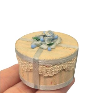 May include: A small round box with a light beige striped pattern and a floral print. The lid is decorated with a light blue fabric rose and small blue beads. A cream-colored lace trim encircles the top edge, and a light blue ribbon crosses the lid.