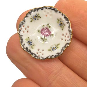 May include: A miniature white porcelain plate with a scalloped edge and gold trim. The plate features a hand-painted design of a pink rose in the center, surrounded by small blue and green floral accents. The plate has small holes around the edge.