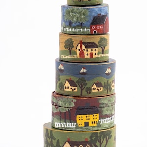 May include: A stack of six round, hand-painted wooden boxes. Each box features a different rural landscape scene with houses, trees, and boats. The color palette includes greens, blues, yellows, and reds, creating a folk art aesthetic.