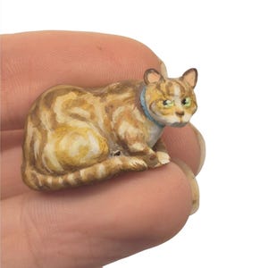 May include: A small, hand-painted figurine of a ginger tabby cat in a resting position. The cat features brown and cream swirled fur patterns, green eyes, and a solid light blue collar around its neck, captured in a detailed miniature form.