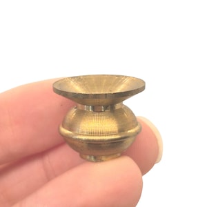 May include: A small, antique brass-colored decorative object with a round base and a flared top. The object has a textured surface and appears to be made of metal. The object is held in a hand against a white background.
