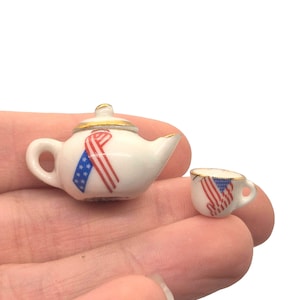 May include: Miniature white porcelain teapot and teacup set. The teapot has a gold handle and lid, and both pieces are decorated with a red, white, and blue ribbon design. The set is small, perfect for a dollhouse or display.