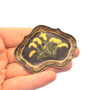 Può includere: Una spilla di forma ovale irregolare caratterizzata da un centro in smalto nero con un disegno dipinto a mano di fiori gialli e fogliame verde. Il pezzo è incorniciato da un bordo decorativo dorato in rilievo con un intricato motivo a filigrana.